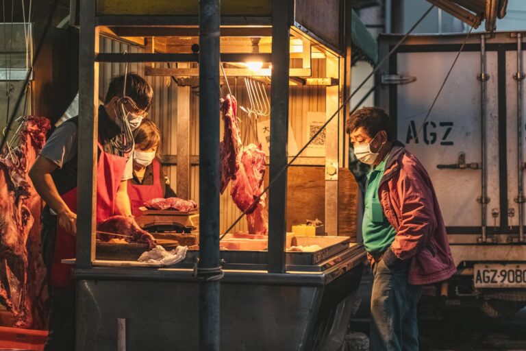 Masked vendors selling fresh meat at an outdoor market stall during nighttime.